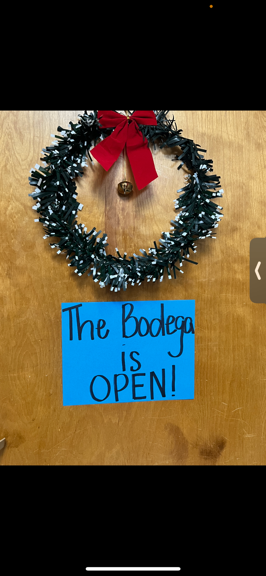 Handmade sign on a wooden door reading 'The Bodega is OPEN!' beneath a holiday wreath