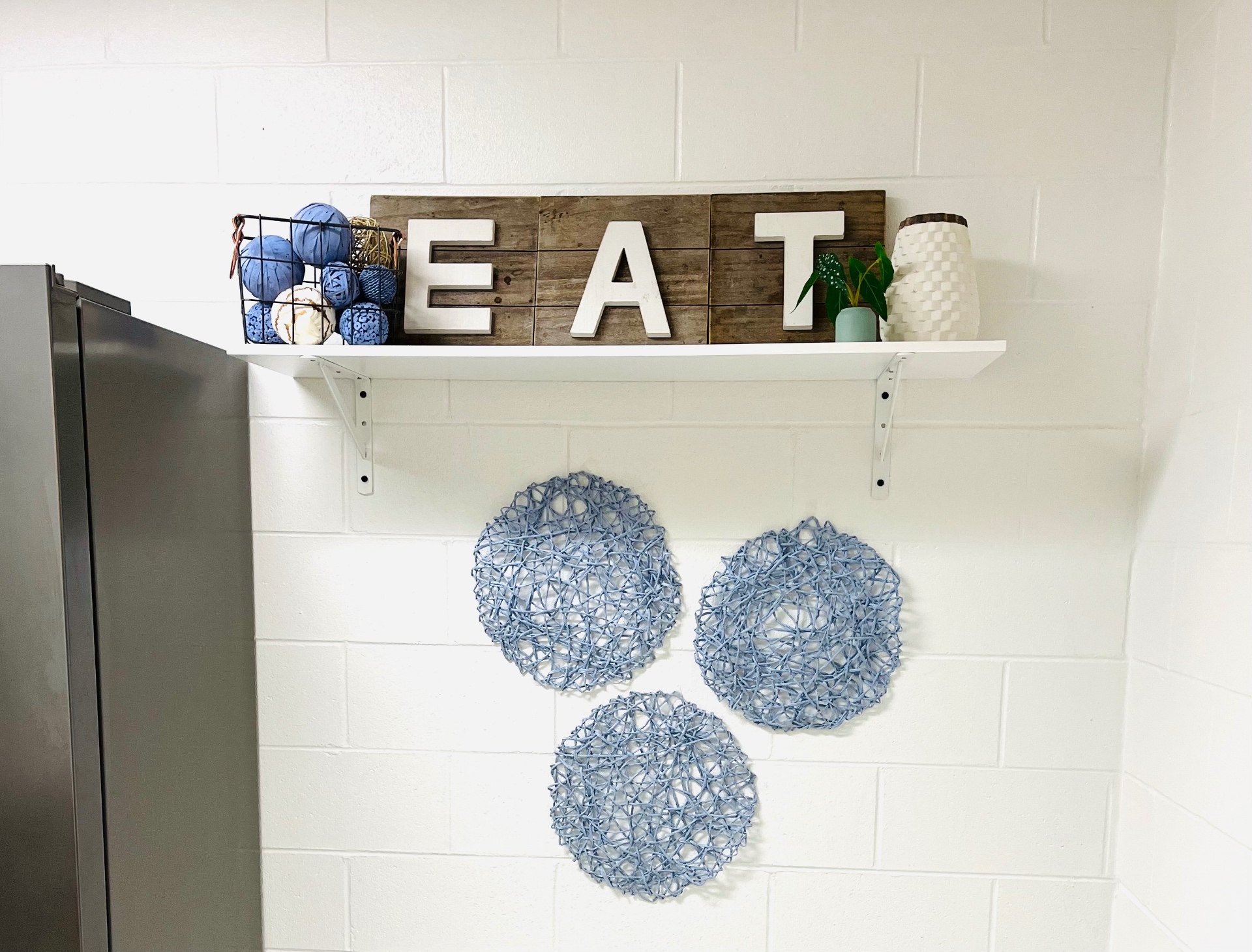 A bright classroom wall with a wooden EAT sign, a basket of soft yarn balls, and woven blue wall decor